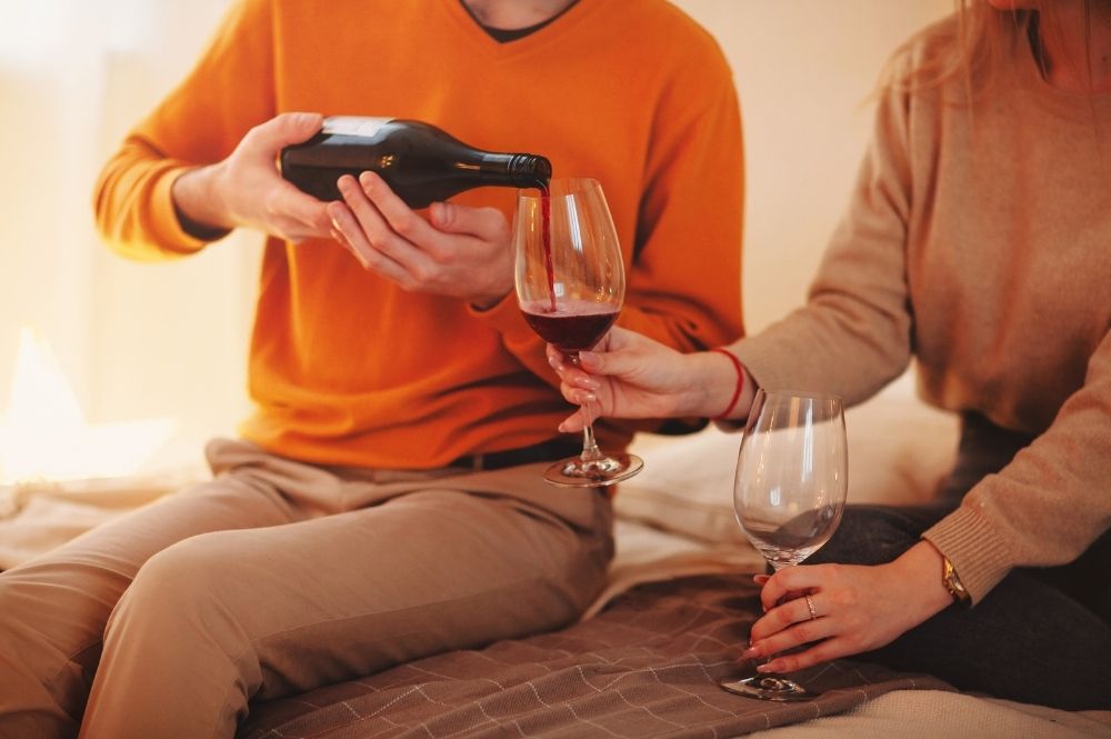 Two people are sitting together with wine glasses, one holds the glasses while the other pours the wine
